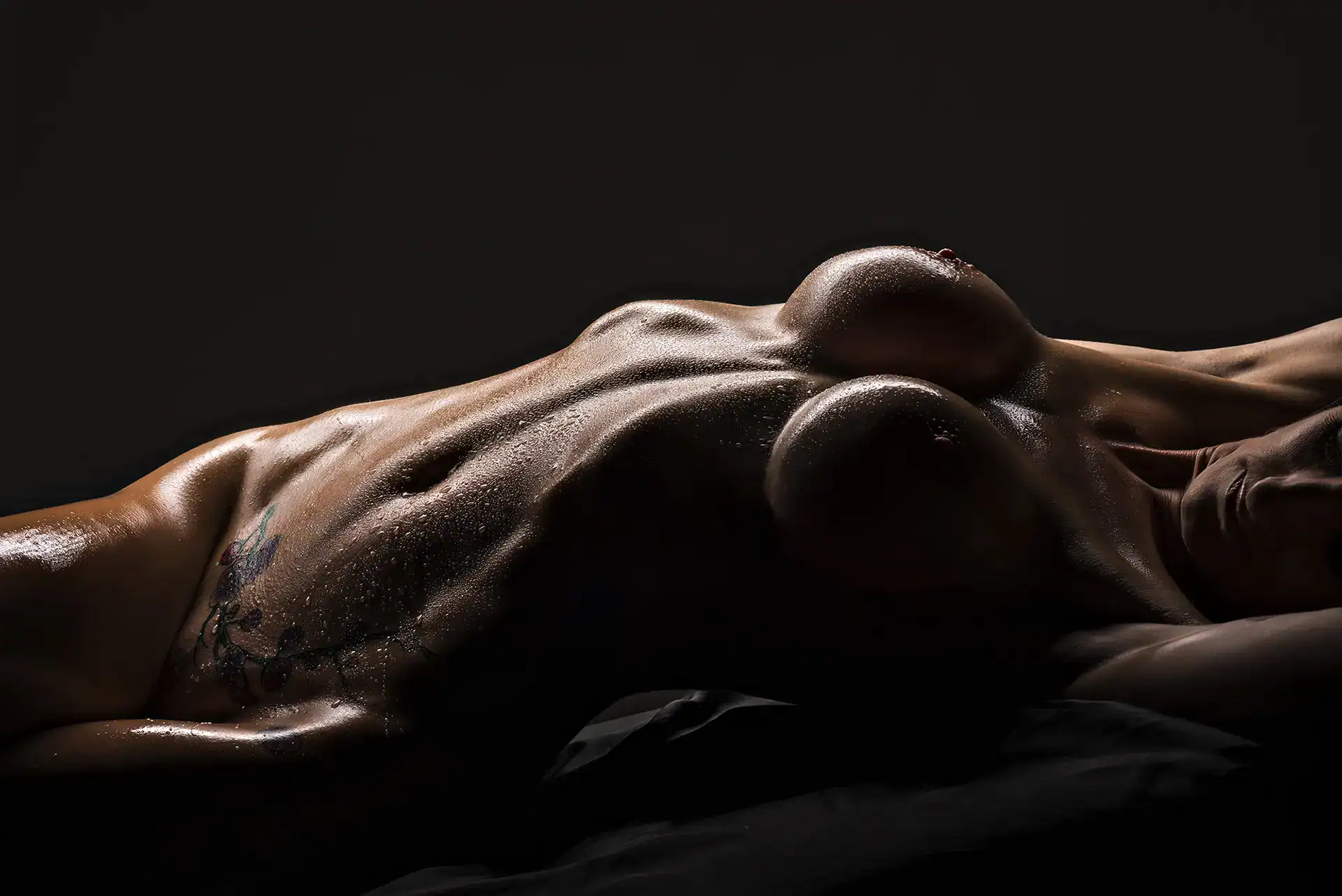Fine art nude of a reclining tattooed woman, her glistening breasts and torso highlighted with water droplets and soft shadow, partially showing her face, Johannesburg studio nude photography