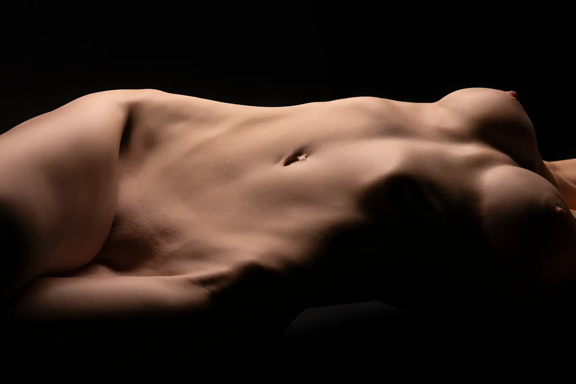 Fine art nude photograph of a reclining woman, highlighting the breasts, waist, and soft shadows along the torso in a warm studio setting, Johannesburg nude photography