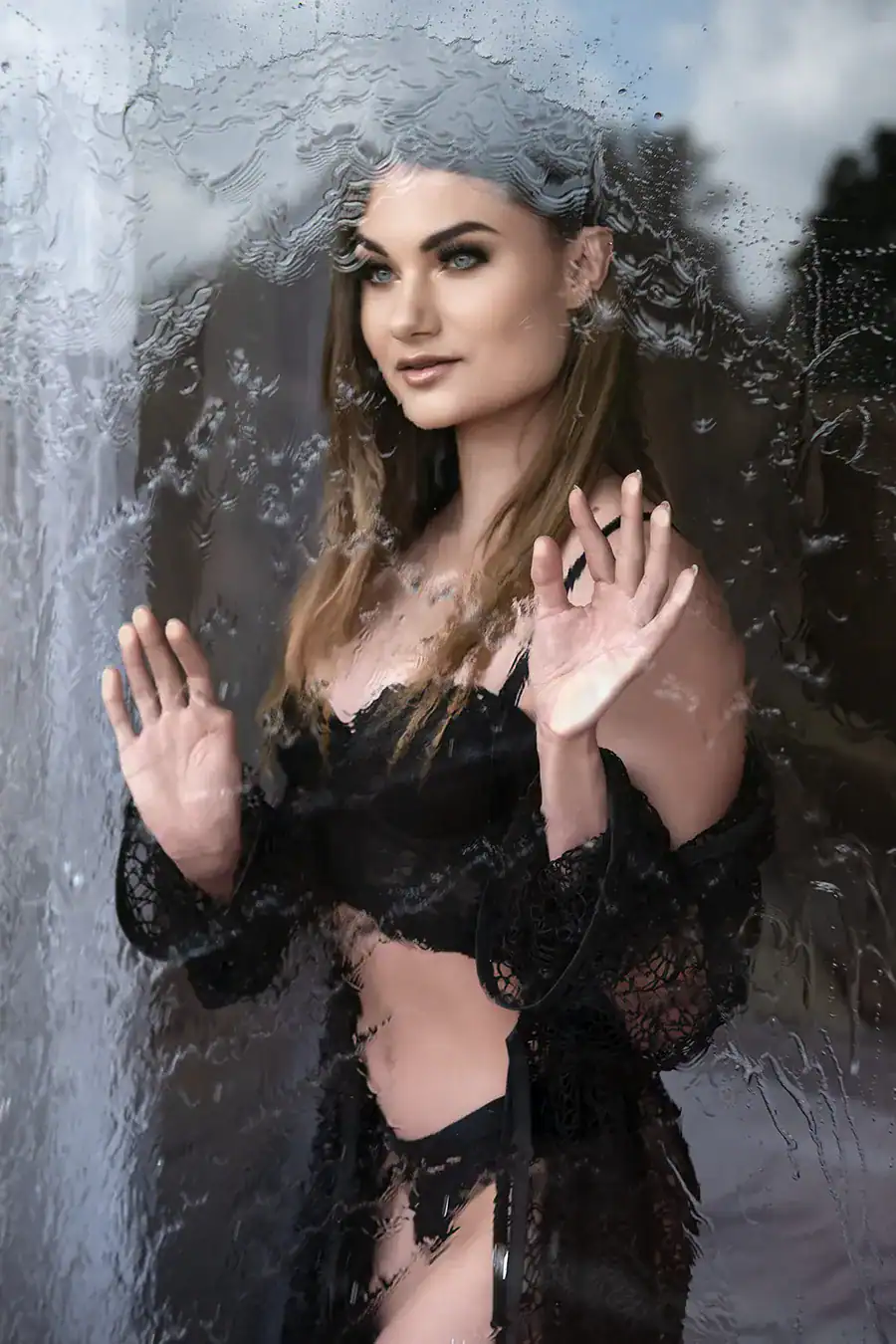 Woman in black lingerie and robe standing behind a rain-covered glass window, captured in a moody and artistic boudoir portrait in Johannesburg