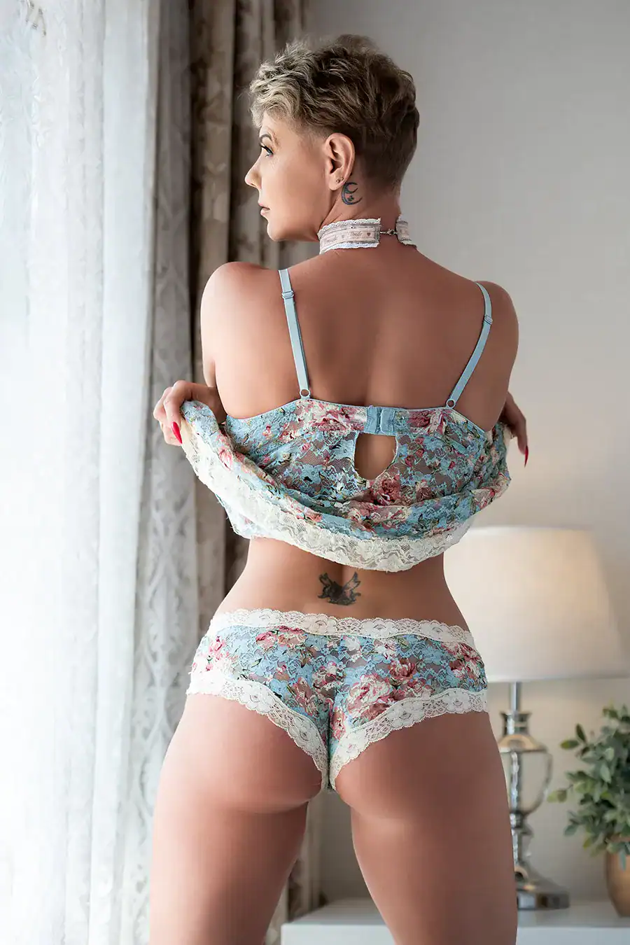 Mature woman in pastel floral lace lingerie facing the window, showing her back and tattoos in a soft-lit boudoir session in Johannesburg