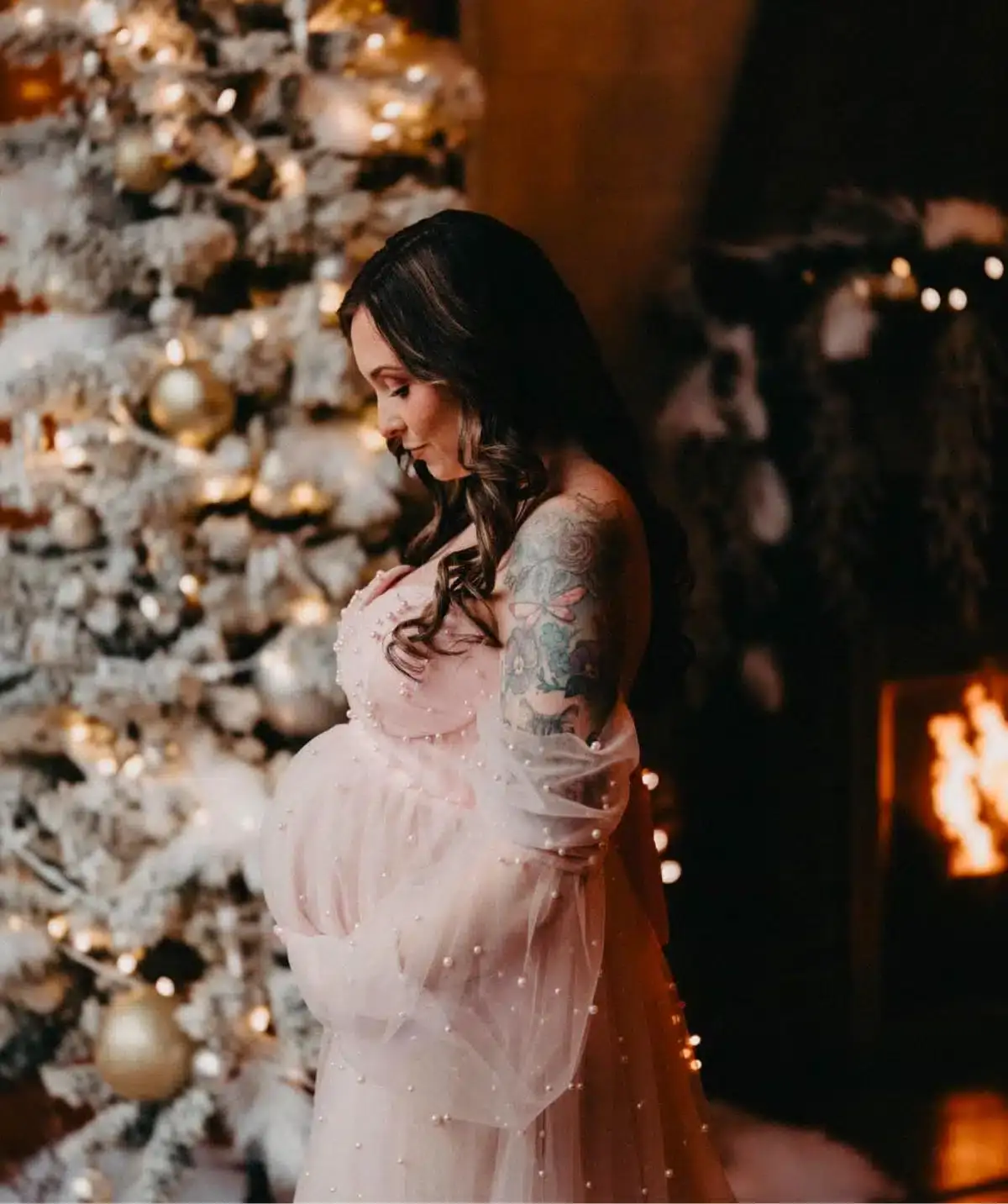 Pregnant woman with dark hair and floral tattoo, standing near a decorated Christmas tree.