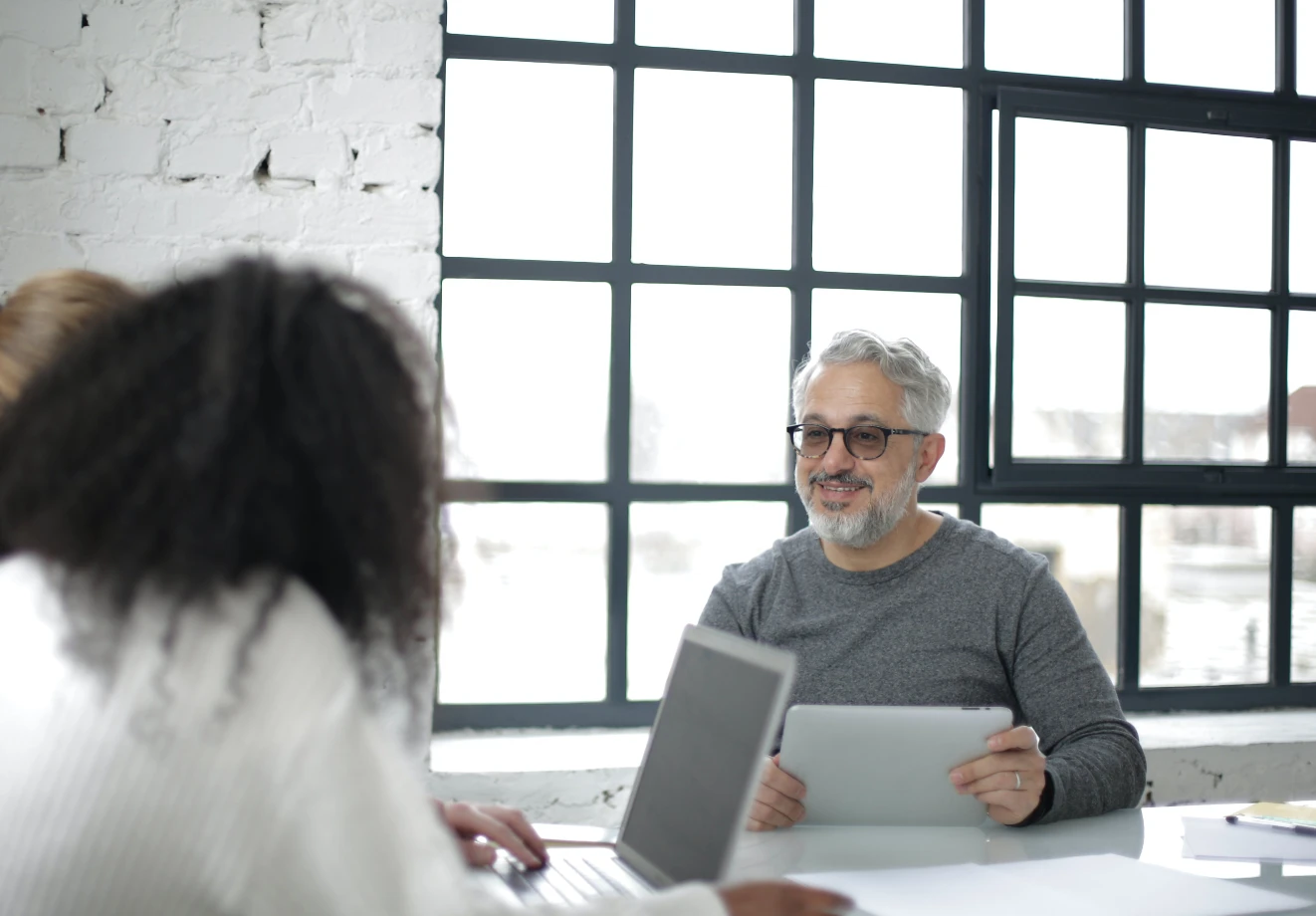 Man sitting at a desk holding a tablet and talking to a colleague across the table from him