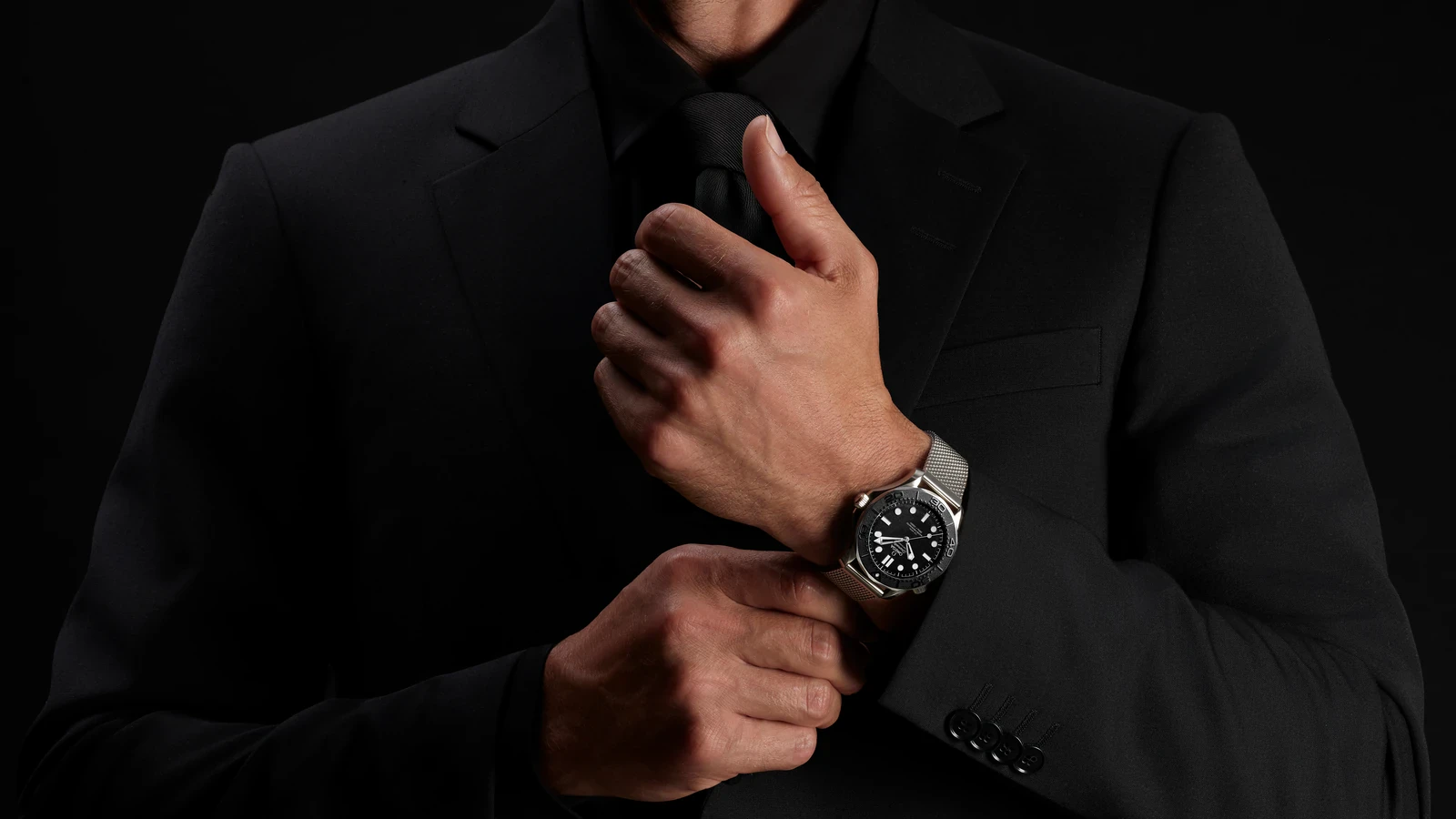 The Spectre edition from the Omega Seamaster James Bond Spectre collection worn on the wrist of a man in a black suit adjusting his cuff, showing its black ceramic bezel, black dial and steel mesh bracelet