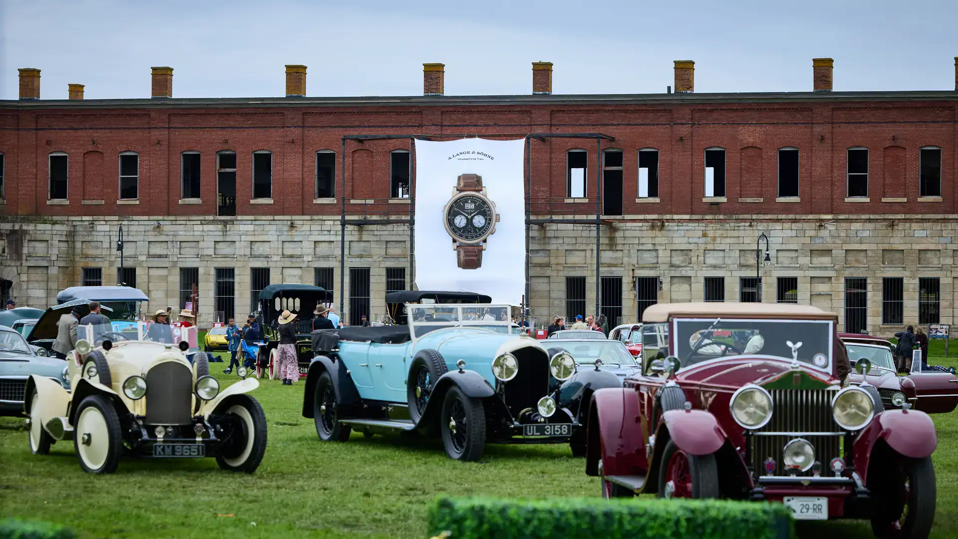 Audrain Newport Concours d'Elegance 2026 vintage cars in cream, pale blue, and maroon on the lawn at Fort Adams in Newport, RI, beneath a large A. Lange & Söhne watch banner