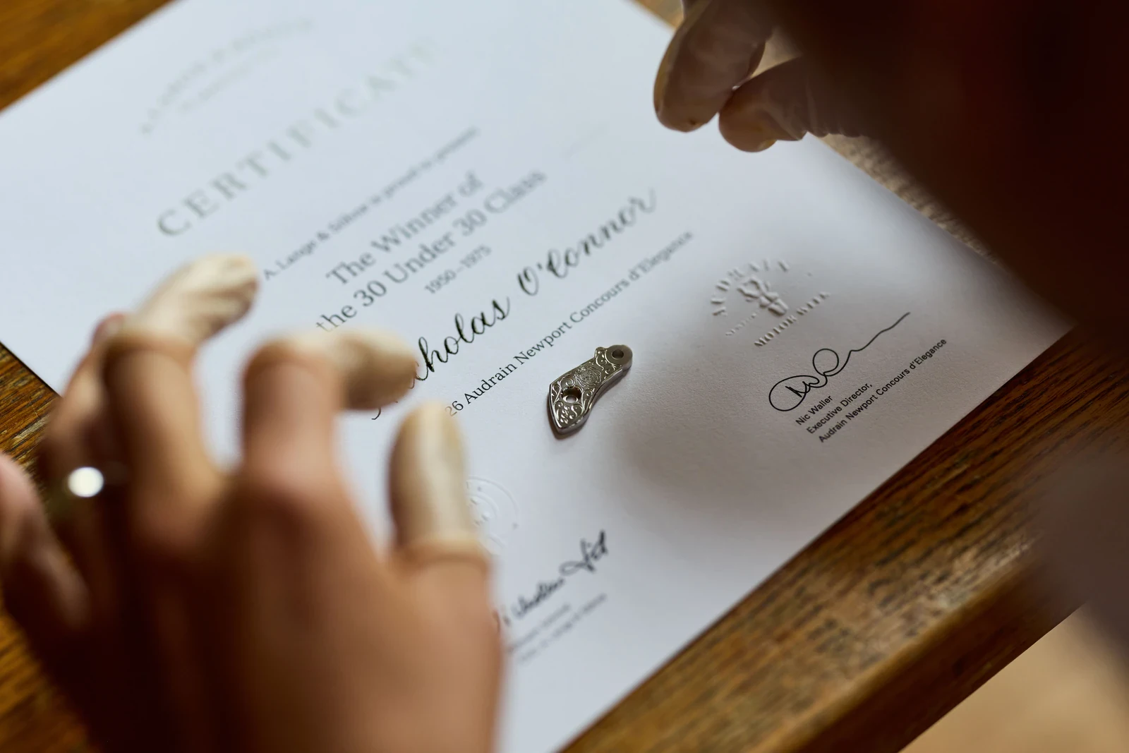 Gloved hands place an engraved balance cock on an A. Lange & Söhne 30 Under 30 class winner's certificate for the Audrain Newport Concours d'Elegance 2026