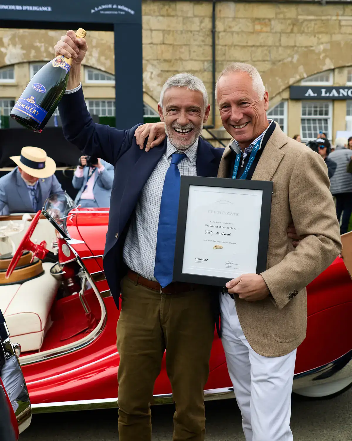 Two men celebrate with champagne and the A. Lange & Söhne Best of Show certificate beside a red open cabriolet at the Audrain Newport Concours d'Elegance 2026
