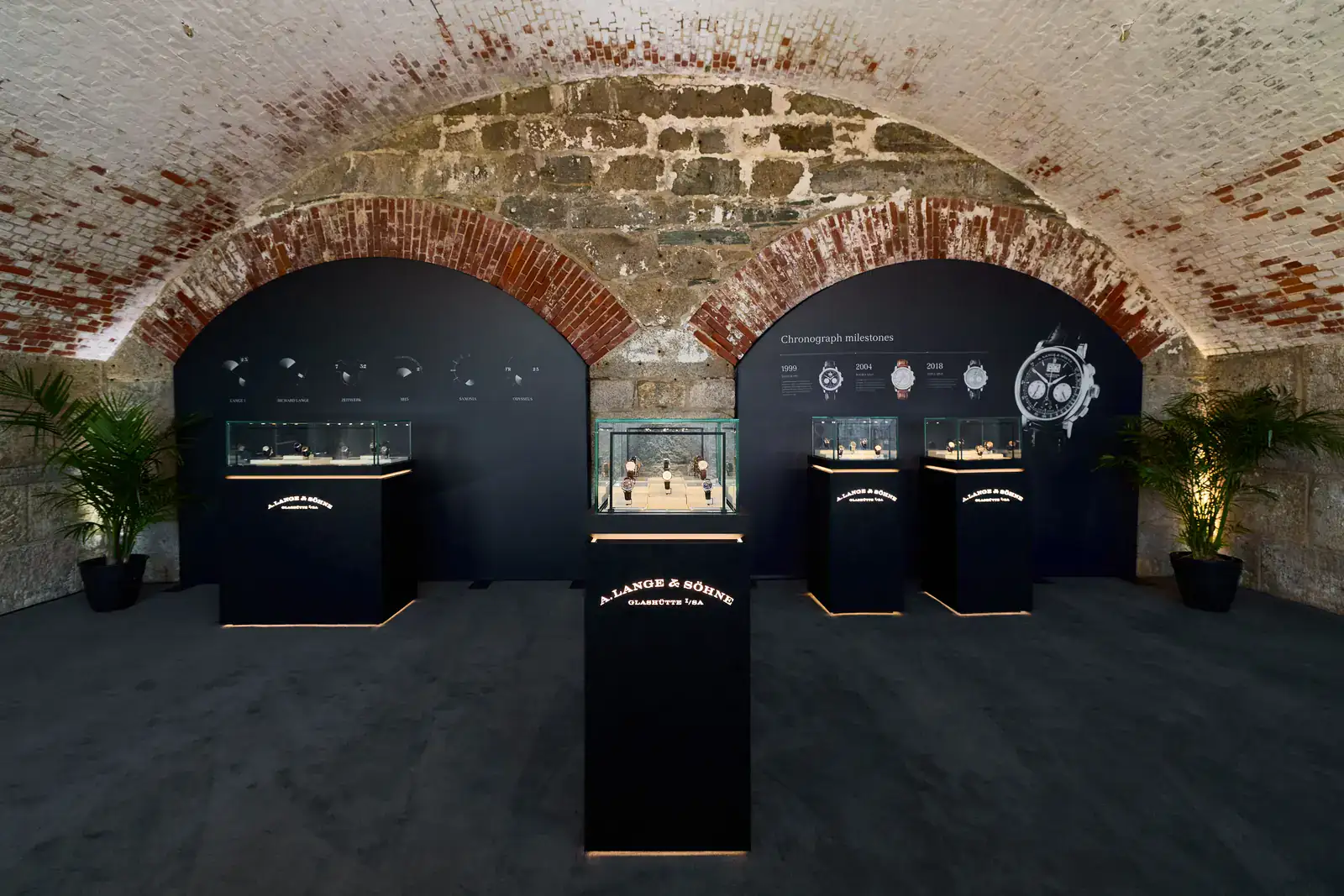 A. Lange & Söhne chronograph gallery at the Audrain Newport Concours d'Elegance 2026, with lit vitrines under brick vaults and a Chronograph milestones wall