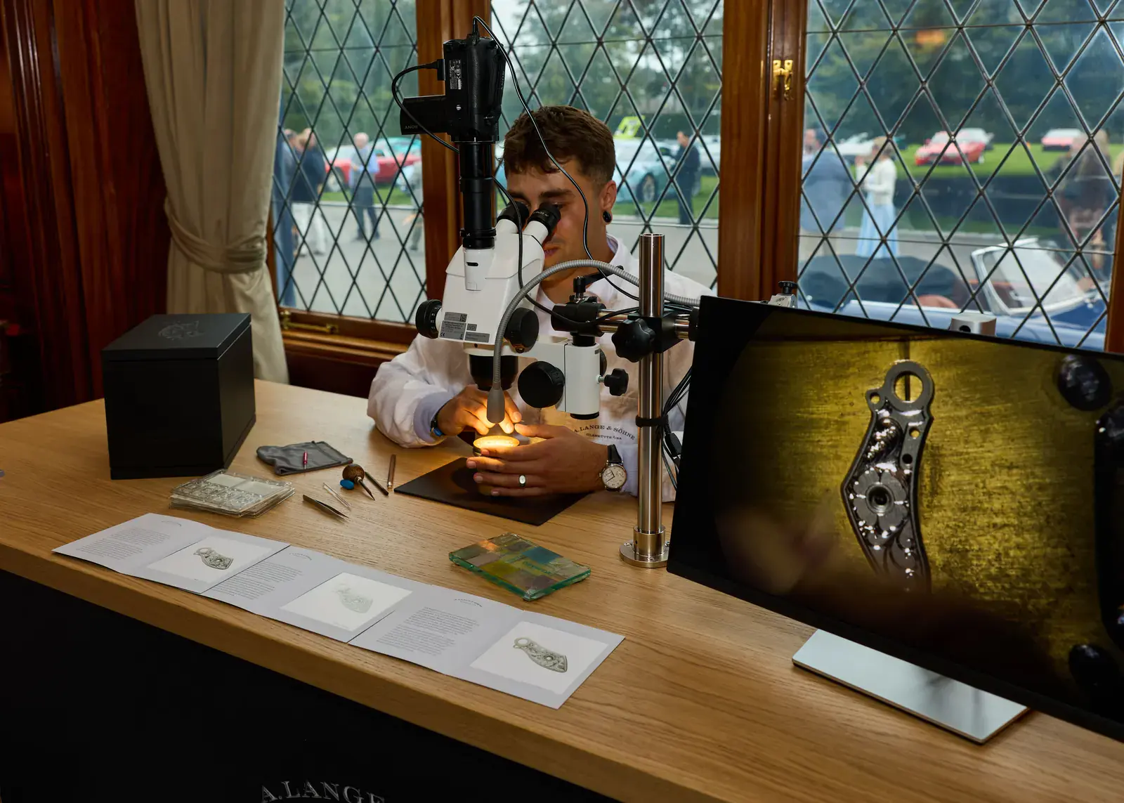 A. Lange & Söhne engraver at a microscope hand-engraving a balance cock, shown magnified on a monitor, during a live demonstration at the Audrain Newport Concours d'Elegance 2026