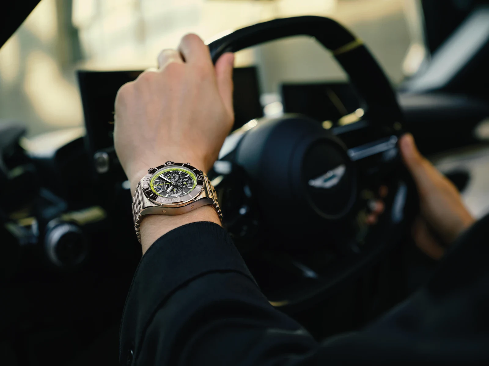 A driver wearing the Breitling Super Chronomat Aston Martin Valhalla B19 44 Perpetual Calendar Chronograph on its Rouleaux bracelet, hands on the steering wheel