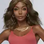 Elegant woman with wavy, light brown wig, wearing coral top and jewelry.