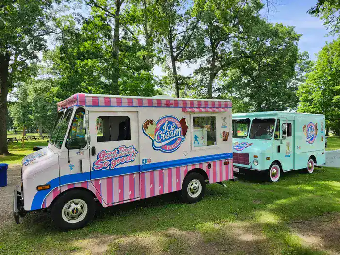 Two colorful ice cream trucks parked outdoors with trees in the background.