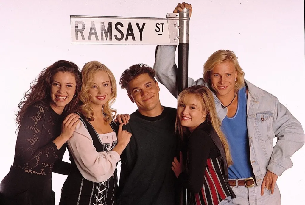 Four young adults (three women and two men) wearing 1900s era clothes and hairstyles, smiling under a 'Ramsay St' street sign.