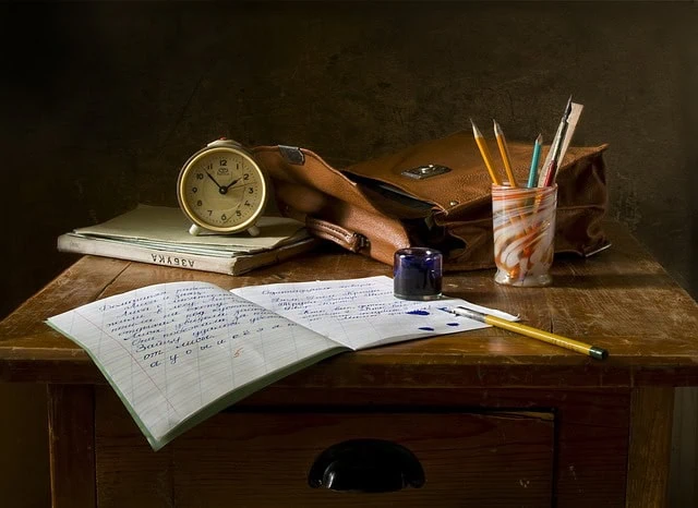 A clock, notebooks, pencils, and a pen on a wooden desk with a bag, creating a study or workspace scene.