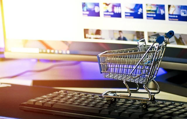 Shopping cart model placed on computer keyboard with monitor displaying online shopping website in the background; digital commerce concept.