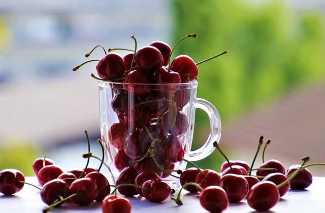 Fresh ripe cherries in a glass mug with some spilled on a white surface, showcasing natural fruit selection for tea ingredients or snacks, with a blurred green outdoor background.