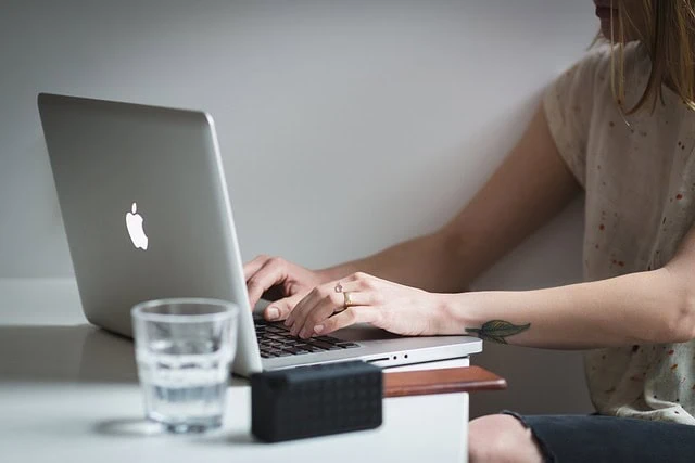Laptop with Apple logo on a white desk, woman working on her computer, glass of water and portable speaker nearby.