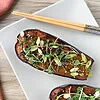 Moitié d’aubergine laquée au miso, parsemée de ciboule, de pousses et de graines de sésame, dans une assiette blanche avec des baguettes