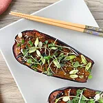 Moitié d’aubergine laquée au miso, parsemée de ciboule, de pousses et de graines de sésame, dans une assiette blanche avec des baguettes