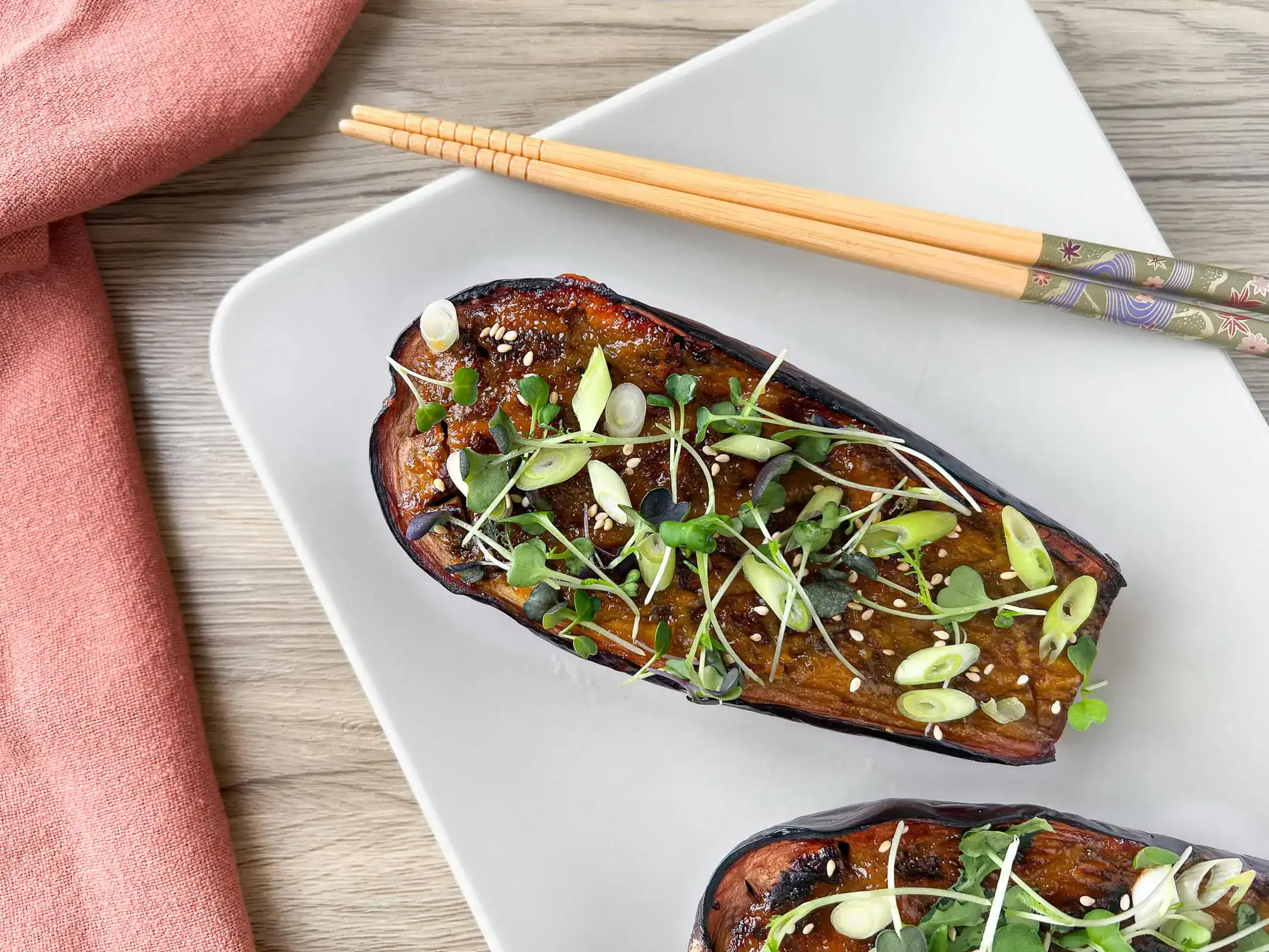 Moitié d’aubergine laquée au miso, parsemée de ciboule, de pousses et de graines de sésame, dans une assiette blanche avec des baguettes