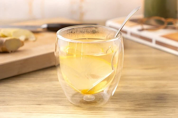 Ginger lemon honey tea in a double-walled glass, with a lemon slice and a spoon, on a light wood countertop