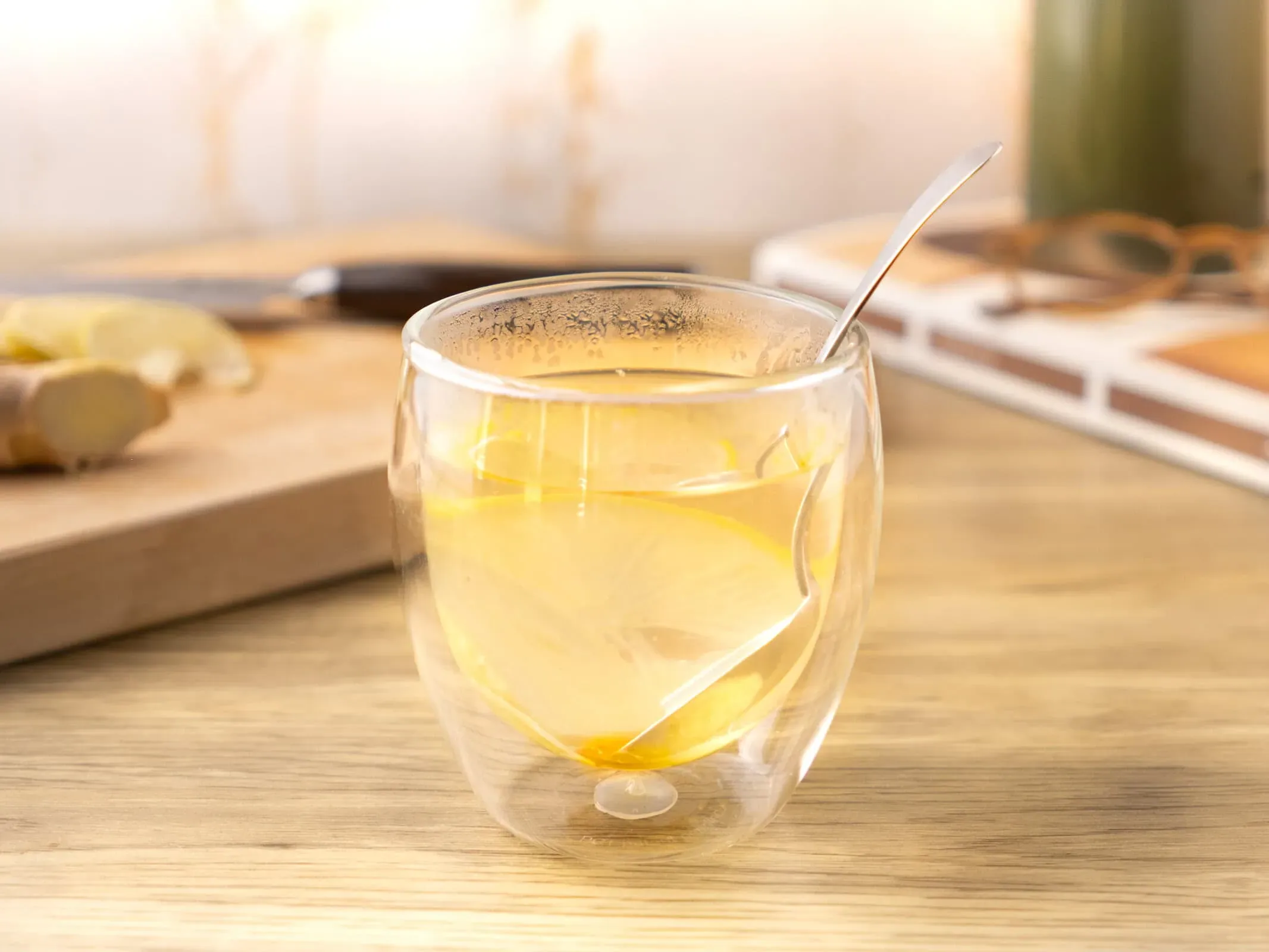 Infusion gingembre citron dans un verre à double paroi, avec une rondelle de citron et une cuillère, sur un plan de travail en bois clair