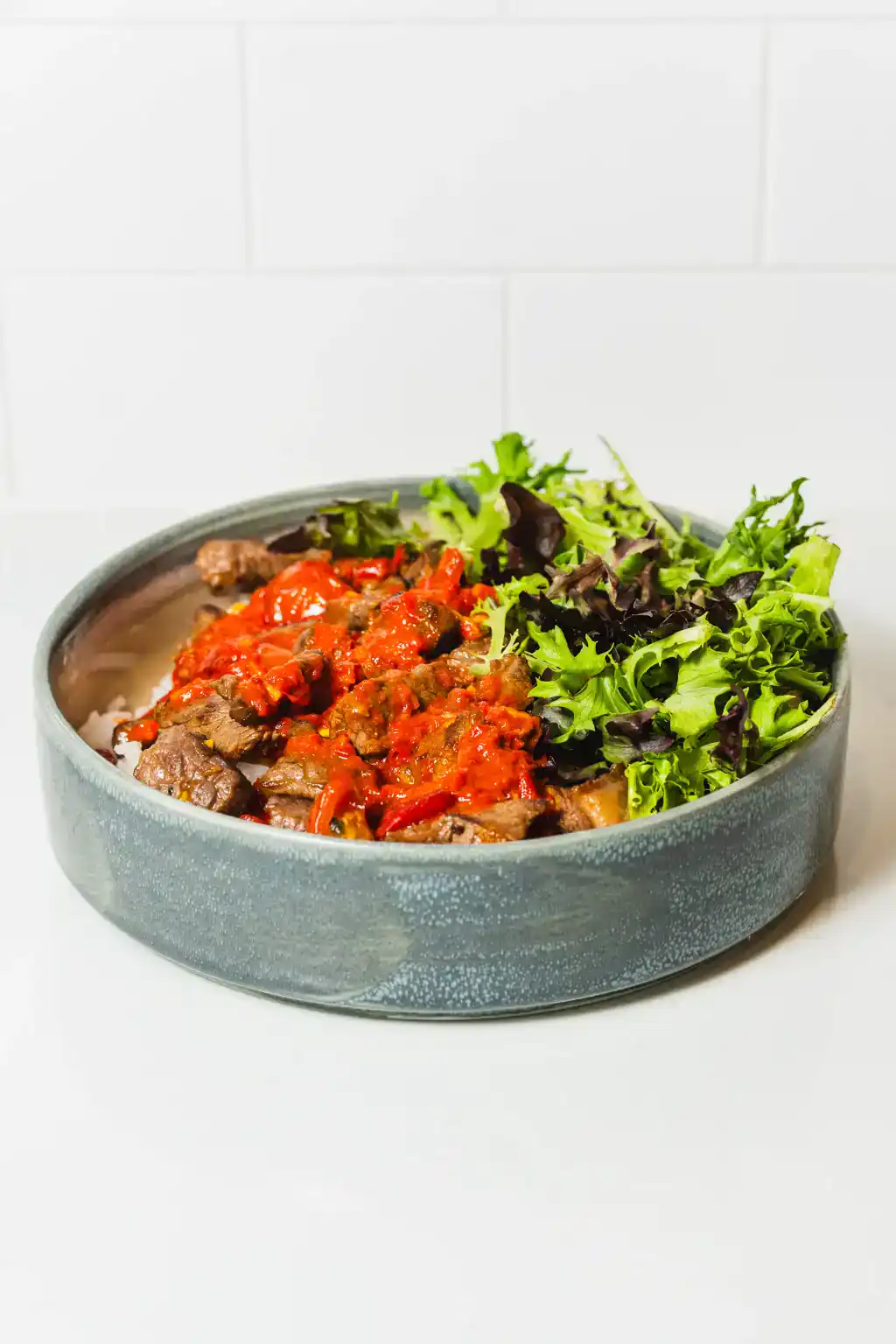Steak bowl with cooked beef, tomato sauce, and fresh leafy greens in a ceramic dish.