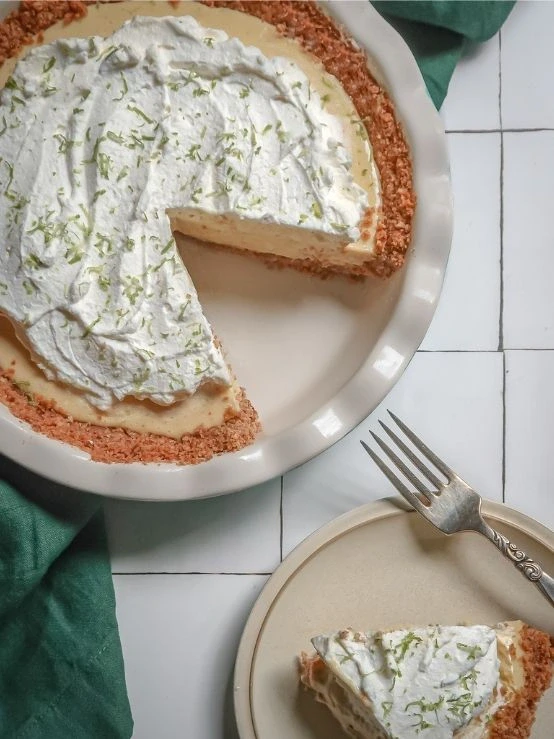 Key lime pie with pecan graham cracker crust topped with whipped cream and lime zest, with a slice served on a plate.