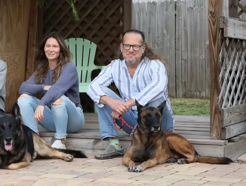 Two people sitting on a porch with two German shepherd dogs, sharing testimonials about dog training.