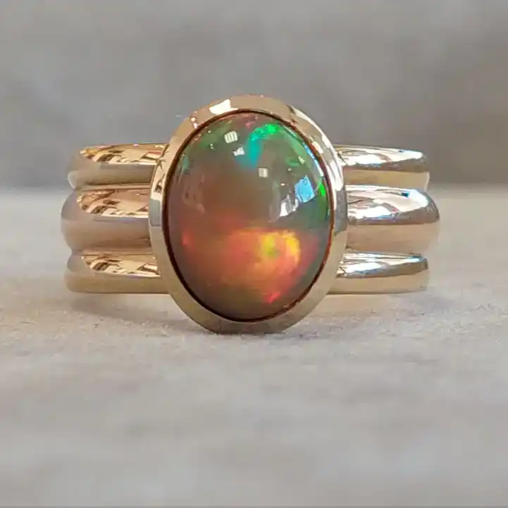 Goldring mit einem ovalen Opal, der grüne und rote Farbreflexe zeigt, auf einem hellen Untergrund.