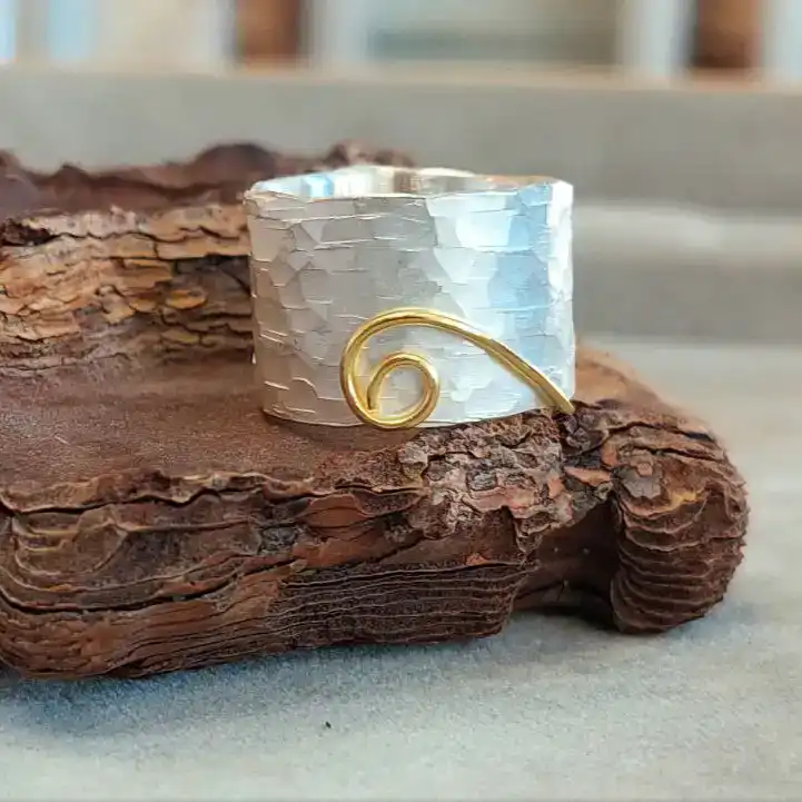 Silberner Ring mit gehämmertem Design und einer goldenen Spirale, auf einer Holzoberfläche platziert.