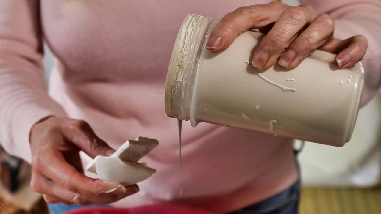 A person in a pink sweater is pouring creamy liquid from a jar onto a piece of a broken mug.