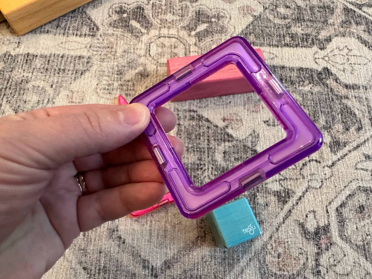 A woman's hand holds a purple square Magformer with 4 magnets visible, one on each side. There are some Tegu blocks in the background.