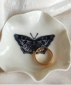 The Butterfly Ring Holder, a white ceramic dish featuring a black butterfly design, holds a single gold ring.