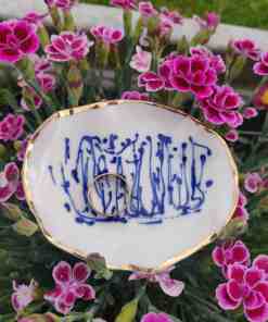 A Blue and White Porcelain Ring Holder with gold lustre is surrounded by vibrant pink flowers, showcasing its small white dish adorned with intricate blue patterns.