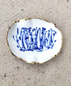 The Blue and White Porcelain Ring Holder with gold lustre features a white base adorned with abstract blue patterns and is elegantly finished with gold-trimmed edges, all set against a textured gray surface.
