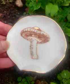 A close-up of the Enchanted Forest Porcelain Mushroom Ring Holder, showcasing its detailed mushroom illustration and gold lustre, held by a hand against a backdrop of green leaves.