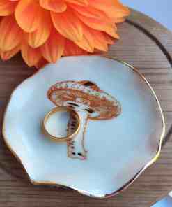 A gold ring is elegantly displayed on the Enchanted Forest Porcelain Mushroom Ring Holder/Trinket with Gold Lustre. The holder is set on a wooden surface beside an orange flower.