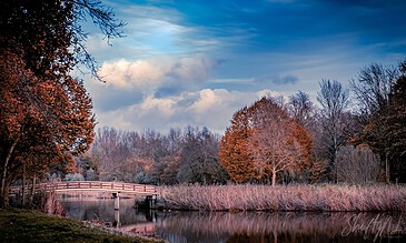 Serene autumn landscape featuring a wooden bridge over a calm river, surrounded by colorful fall foliage and reflected in the water, under a partly cloudy sky.