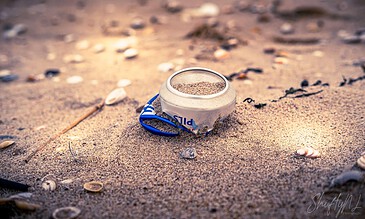 A discarded beer can partially buried in sandy beach with seashells scattered around, highlighting pollution in natural coastal environments.