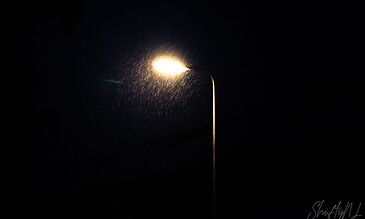A lone streetlight shining through the rain at night, creating a dramatic and moody atmosphere with illuminated raindrops against a dark sky.