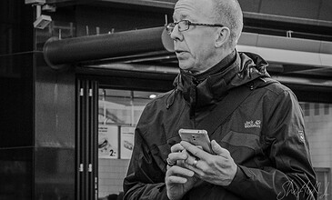 A man with glasses and a jacket looking at his phone in an urban setting, black and white photography capturing a candid moment.