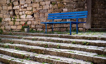A vibrant blue park bench on ancient stone steps next to a weathered stone wall with a pot of red flowers nearby.