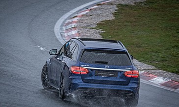 A sleek blue Mercedes-Benz C63 S AMG driving through a wet race track curve in rainy conditions, showcasing dynamic motion and precision driving skills.
