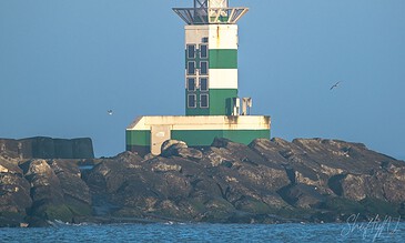 A lighthouse with green and white stripes standing on rocky shoreline amid blue sea and sky.