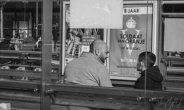 A man and boy sitting on a bus stop bench, engaged in conversation, with posters and reflections in the background, capturing candid moments on city streets.