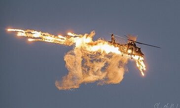 Dynamic image of a helicopter in action during a firefighting operation, with flames and smoke trailing behind. Aerial firefighting helicopter releasing fire retardant with flames and smoke against a clear blue sky.
