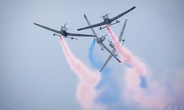 Spectacular aviation display featuring three planes creating vibrant smoke patterns during an airshow. Aerial view of three aircraft performing a synchronized stunt with colorful smoke trails in pink and blue against a cloudy sky.