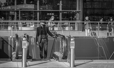A stylish man with a beard wearing a blazer and cap, holding a water bottle, standing at an escalator entrance in front of a modern glass building with people walking by.