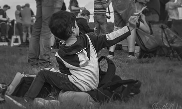 A young boy at an outdoor gathering, sitting on the grass with a backpack, engaging with something in the distance while others stand around him. A boy sitting on the grass at an outdoor event, reaching out with his arm, surrounded by a crowd of people.