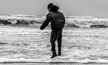 Capturing raw emotion and natural beauty, this black-and-white photography showcases an individual's peaceful moment by the ocean, emphasizing the serenity of the seaside. A young man in a black jacket and pants walks along the shoreline of the beach with waves crashing behind him, capturing a moment of solitude and reflection by the sea.